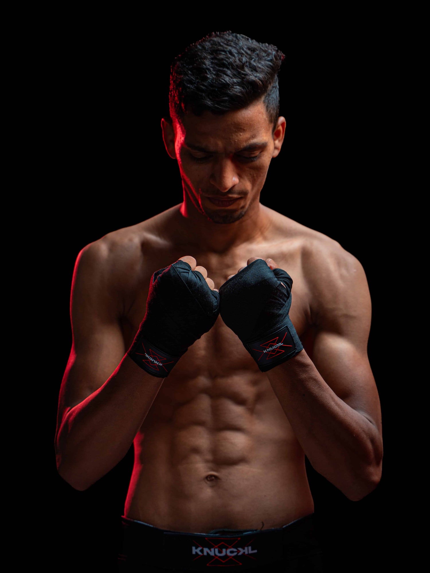 Male athlete wearing Knuckl hand wraps and MMA shorts standing in a focused pose against a black background