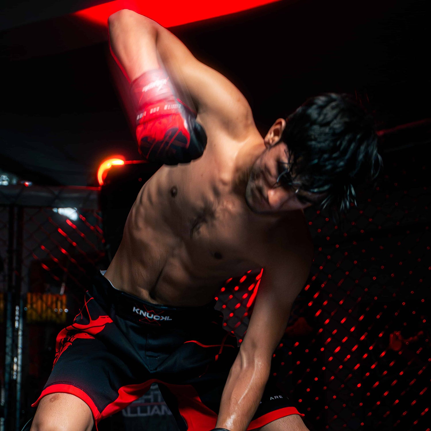 Athlete wearing full Knuckl MMA kit striking a punching bag under red lights in a cage gym.
