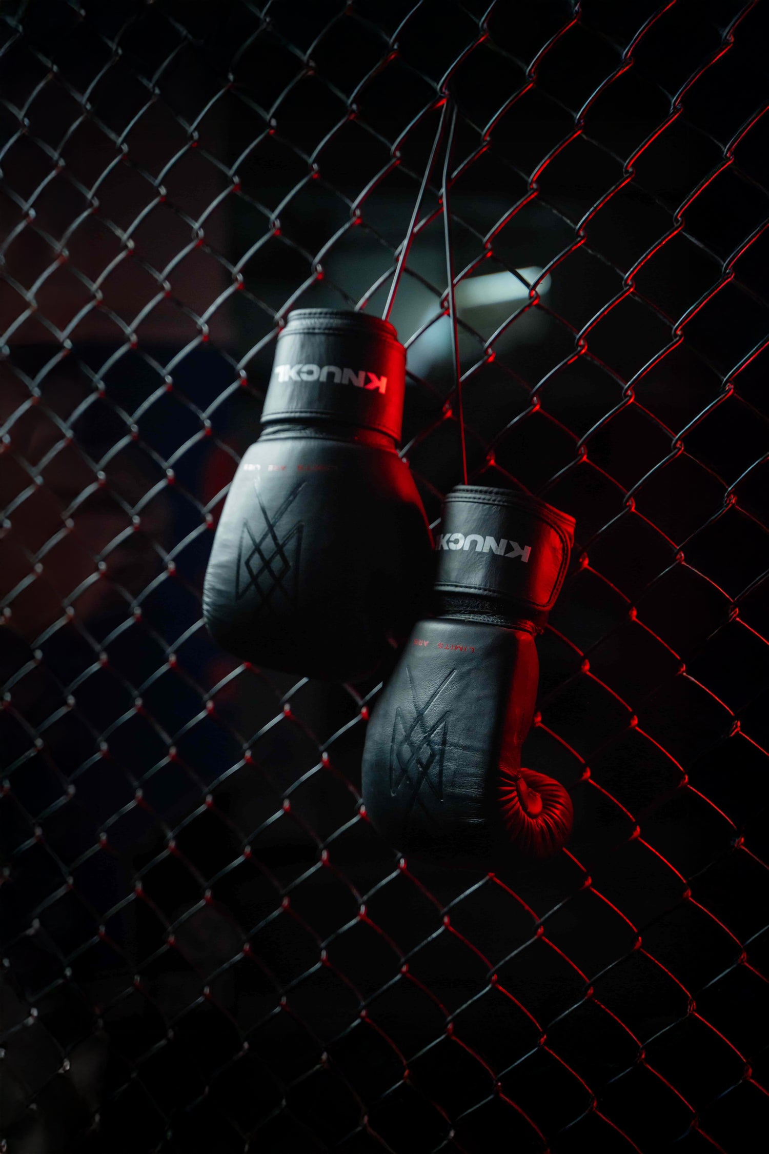 Black leather boxing gloves with embossed Knuckl symbol hanging on chain-link fence under red light