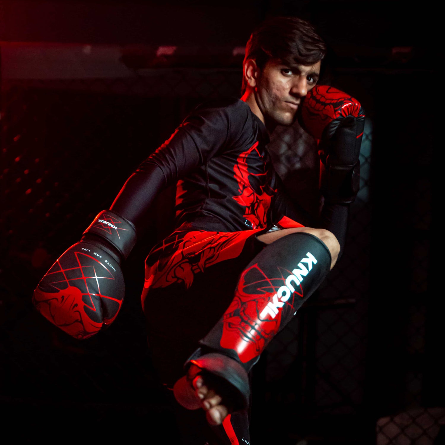 Male fighter wearing full Knuckl MMA gear in high kick pose with red and black gloves, shin guards, and rash guard